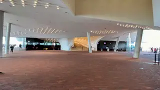 A spacious modern lobby with a brick floor, angled columns, ceiling lights, and a central staircase has a few distant visitors.