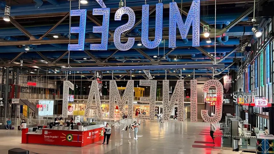 Oversized illuminated letters hang upside down in a spacious transit hall with ticket counters, stairs, and scattered visitors.