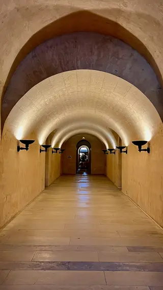 A warmly lit stone corridor with a series of rounded vaulted arches leads to a distant doorway.