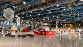 A spacious modern shopping interior with blue exposed ceiling beams, a red information counter, and scattered visitors.