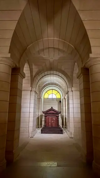 A vaulted stone corridor with arched columns leads to an ornate wooden structure beneath a yellow stained-glass window.