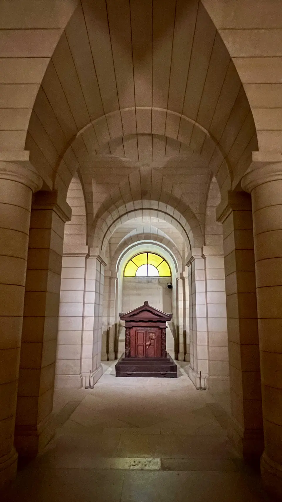 A vaulted stone corridor with arched columns leads to an ornate wooden structure beneath a yellow stained-glass window.