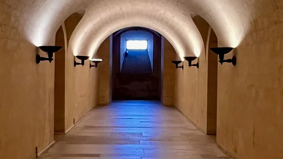 A vaulted stone corridor with wall-mounted lamps leads to a bright window above a short staircase.