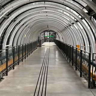 A covered pedestrian walkway with curved metal ribs, glass panels, black railings, and a concrete floor stretches toward a distant entrance.