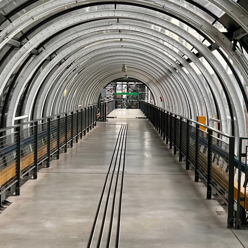 A covered pedestrian walkway with curved metal ribs, glass panels, black railings, and a concrete floor stretches toward a distant entrance.
