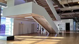 A broad wooden staircase rises through a modern gallery interior with polished floors, exposed ceiling ducts, and elevator doors.