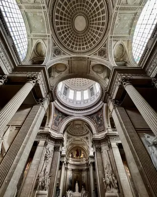 An ornate vaulted interior with coffered ceilings, skylights, and massive fluted columns rises above classical statues.