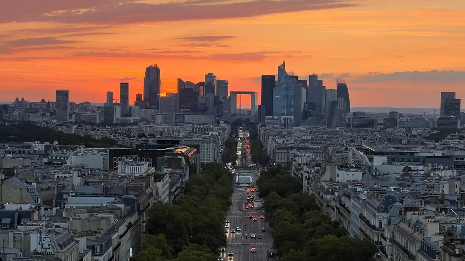 A broad tree-lined avenue leads toward a skyline of high-rise buildings beneath an orange sunset sky.