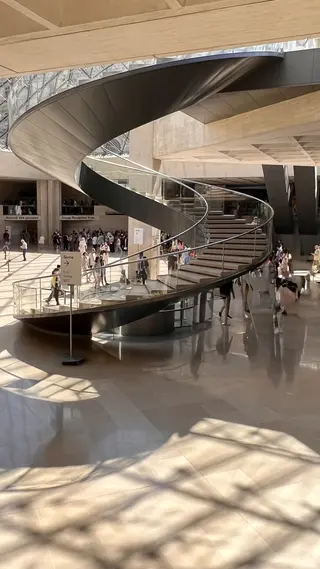 A sweeping black spiral staircase curves through a sunlit atrium with a polished floor and groups of visitors below.