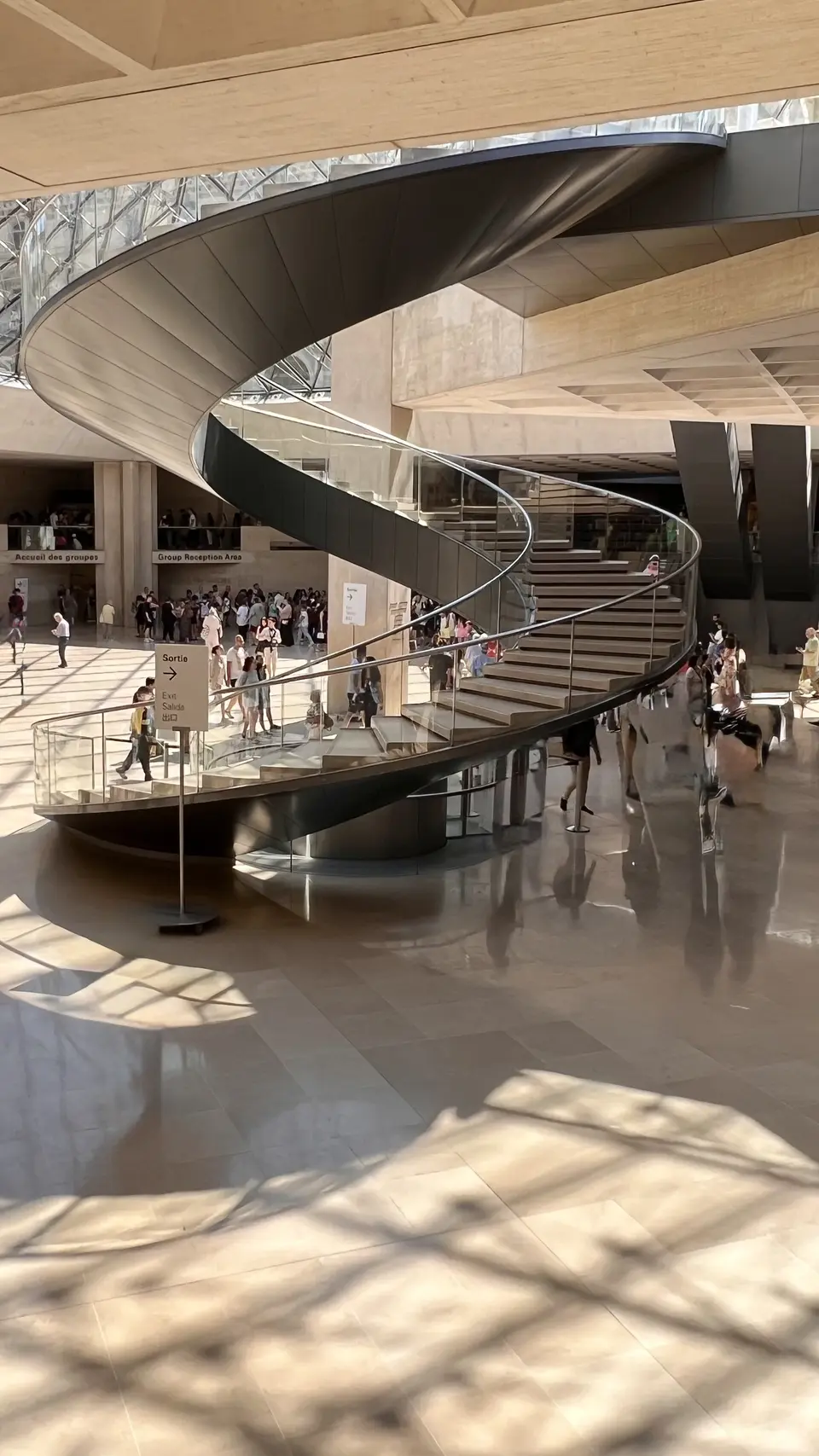 A sweeping black spiral staircase curves through a sunlit atrium with a polished floor and groups of visitors below.