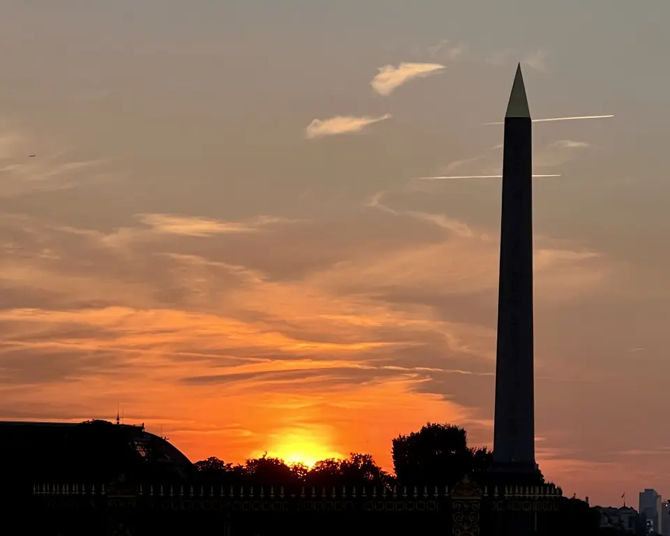 The Washington Monument rises in silhouette against a vivid orange sunset sky above dark treetops and a fence.