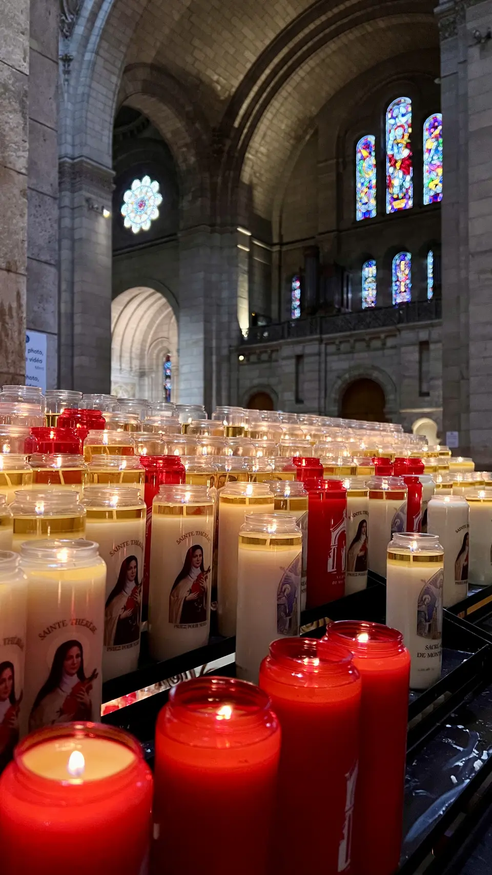 Rows of lit votive candles stand beneath vaulted stone arches and colorful stained-glass windows in a church interior.