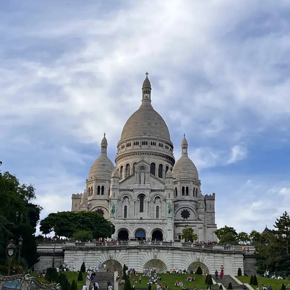 A large white domed church with three towers rises above a terraced lawn beneath a cloudy sky.