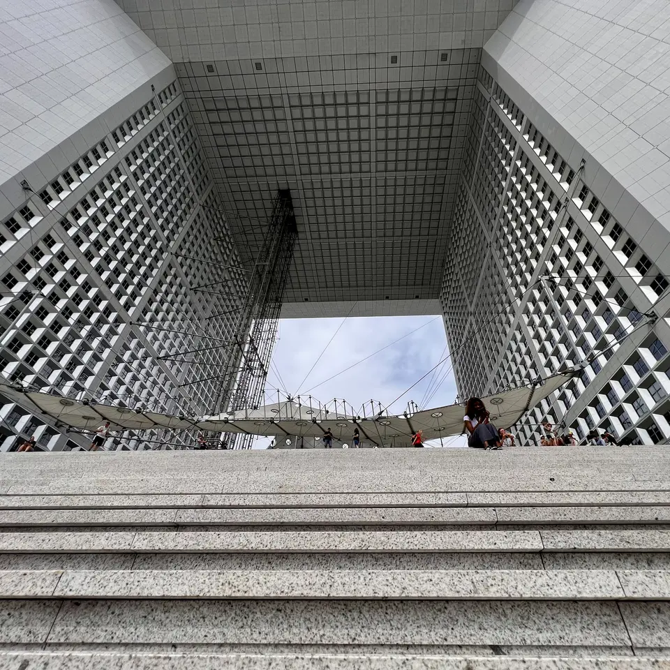 Broad stone steps lead toward the open central arch of a towering grid-windowed building, where people gather beneath a suspended canopy.