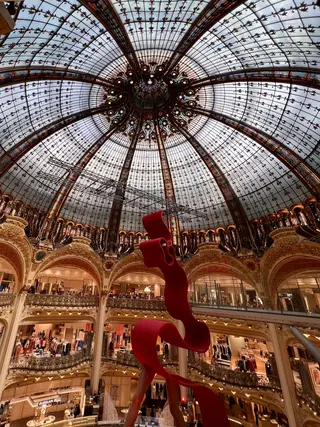 An abstract red hanging sculpture rises beneath an ornate stained-glass dome inside a grand multi-level department store.
