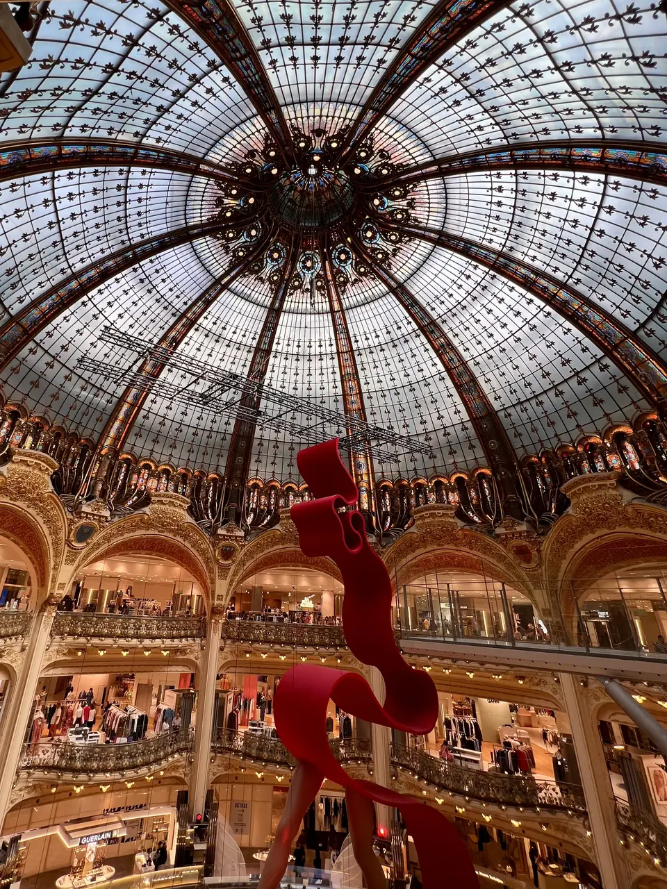 An abstract red hanging sculpture rises beneath an ornate stained-glass dome inside a grand multi-level department store.