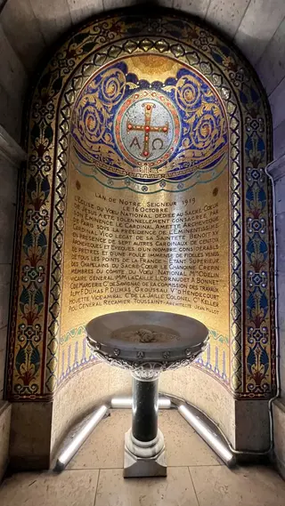 An ornate gold and blue mosaic panel with a cross emblem and French inscription rises behind a decorative stone basin.