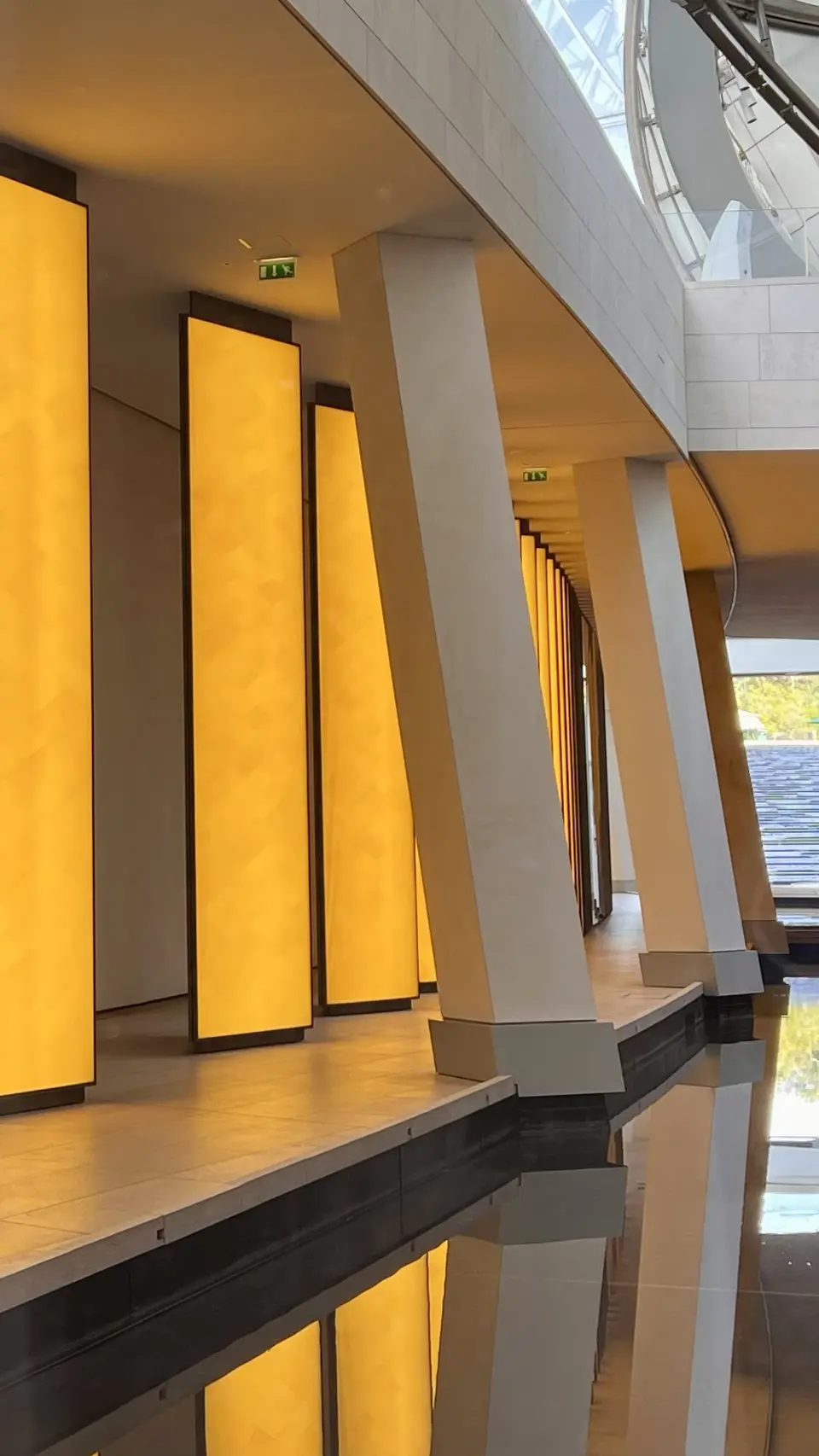 A modern interior corridor features glowing amber wall panels, angled white columns, and reflections in a dark water feature.
