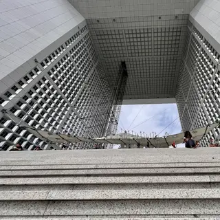 Broad stone steps rise beneath the towering rectangular arch of the Grande Arche de la Défense, with small pedestrians below its gridded facade.