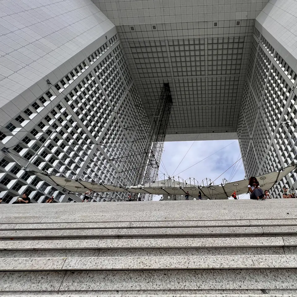 Broad stone steps lead up beneath a monumental open-sided building with gridded windows and a suspended canopy.