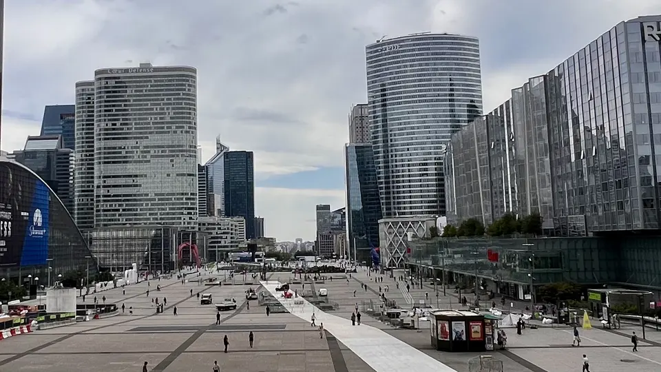 Modern glass-and-steel high-rise buildings surround a broad pedestrian plaza beneath a cloudy sky.
