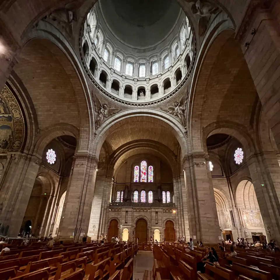 A grand church interior features towering stone arches, a domed ceiling, stained-glass windows, and rows of wooden pews.
