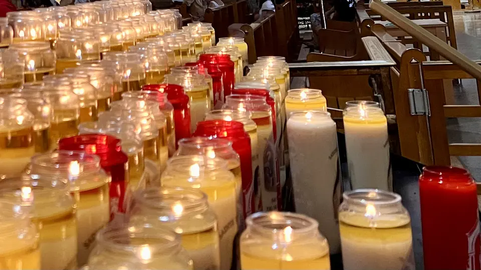 Rows of lit votive candles in glass jars fill a church interior beside wooden pews.