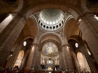 The soaring interior of a stone church features vaulted arches, a bright domed ceiling, stained-glass windows, and a mosaic above the altar.