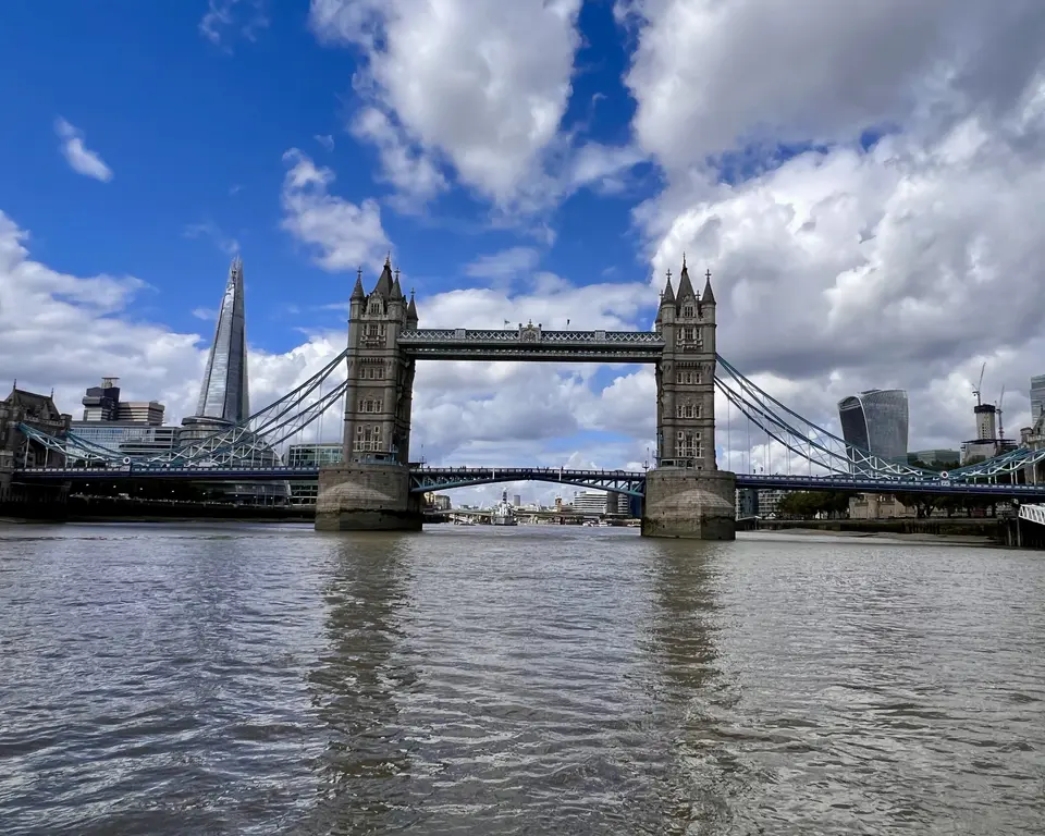 Tower Bridge spans the River Thames beneath a blue sky filled with large clouds, with modern high-rise buildings behind it.