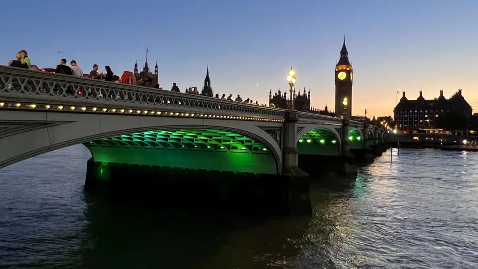 Westminster Bridge, lit green beneath its arches, spans the River Thames at dusk with the illuminated Elizabeth Tower and Houses of Parliament behind it.