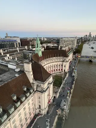 An aerial view shows a grand historic riverside building with a green clock tower beside a broad river, bridge, and modern city skyline.