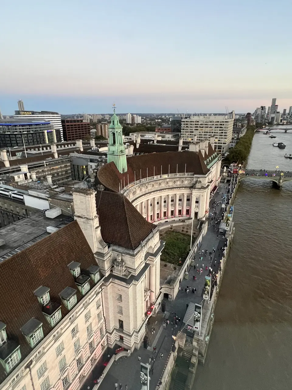An aerial view shows a grand historic riverside building with a green clock tower beside a broad river, bridge, and modern city skyline.