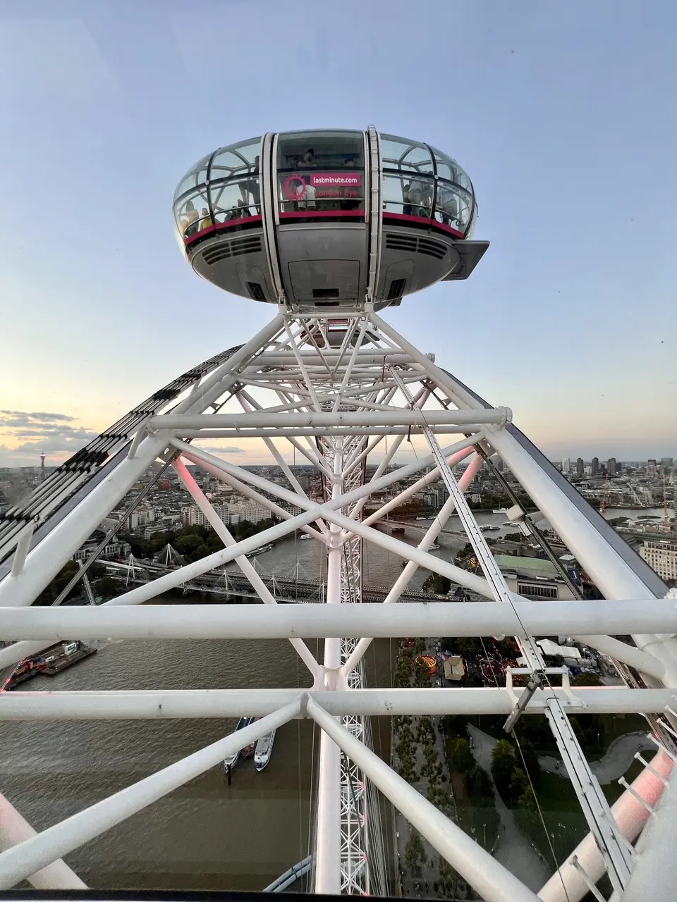 A glass observation capsule bearing a lastminute.com London Eye sign rises above the white framework of a giant wheel, with a river and city skyline below.