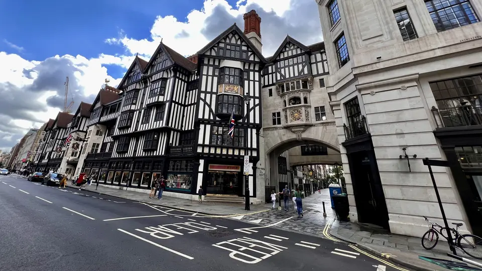 A row of black-and-white timber-framed buildings stands beside a city street, with an ornate stone archway between them under a cloudy blue sky.
