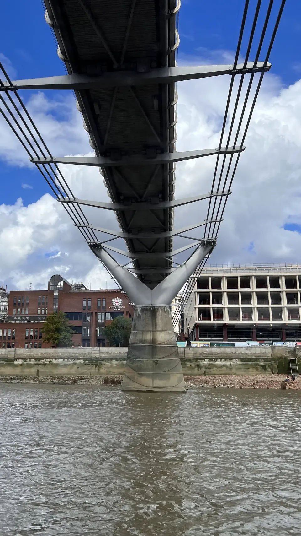 The underside of a cable-supported bridge spans rippling water toward the City of London School and neighboring buildings beneath a blue, cloud-filled sky.