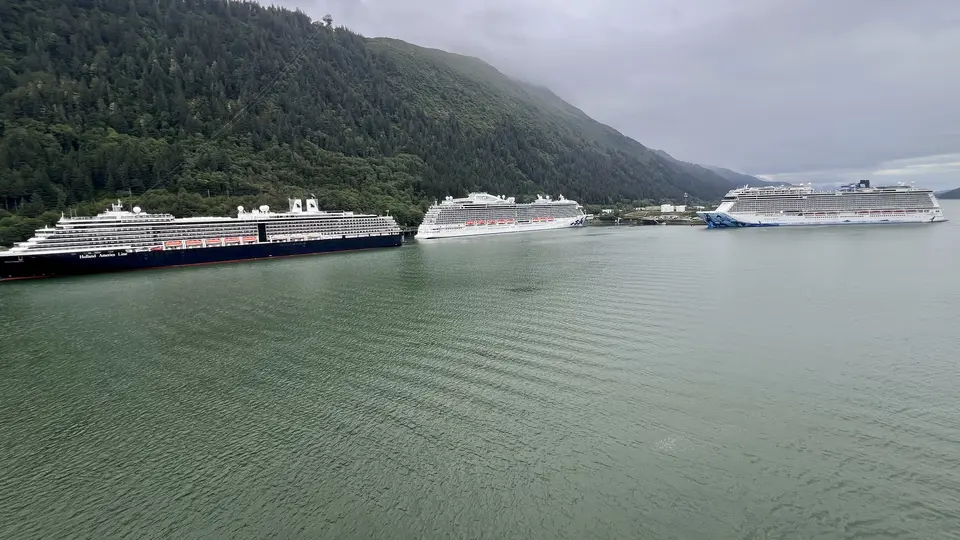 Three large cruise ships are docked along a calm body of water beneath a densely forested mountainside.
