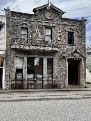 A dark, heavily carved wooden building facade with large decorative letters, curtained windows, and the address number 1849 faces a quiet street.