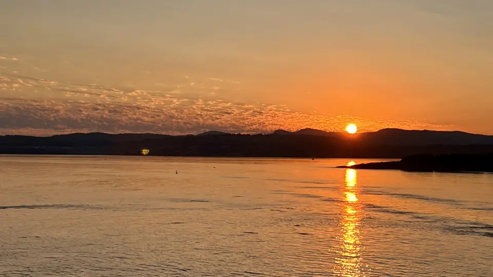 The setting sun casts a bright orange reflection across calm water beneath silhouetted hills.