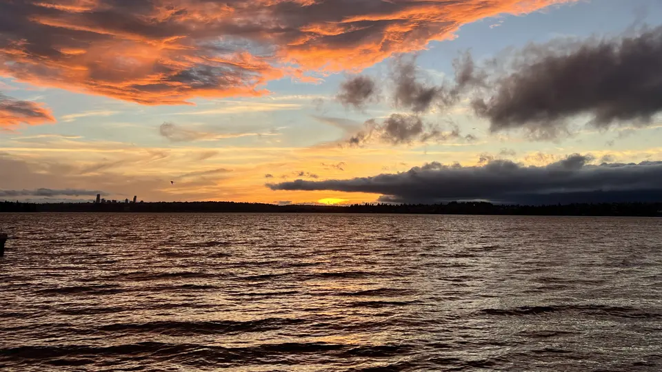 Orange and gray clouds glow above rippling water as the sun sets behind a dark shoreline.