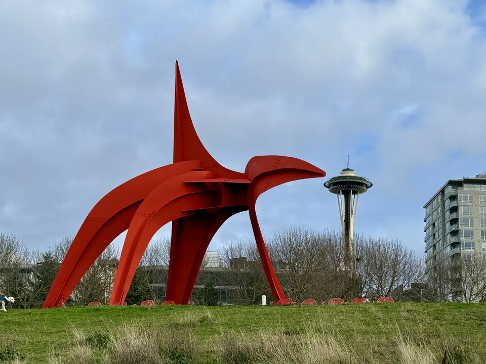 A large red abstract steel sculpture stands on a grassy slope with the Space Needle and nearby buildings behind it.
