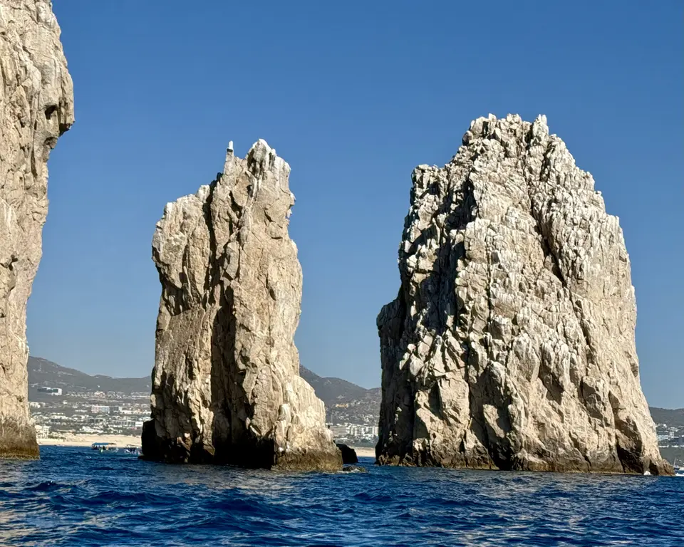 Two towering sea stacks rise from blue water beneath a clear sky, with a coastal town and mountains in the distance.