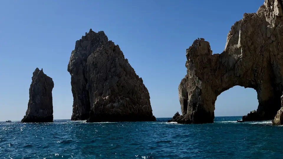 Jagged sea stacks and a natural rock arch rise from the blue ocean beneath a clear sky.