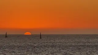 The setting sun glows orange above the ocean horizon as two sailboats cross the rippling water.