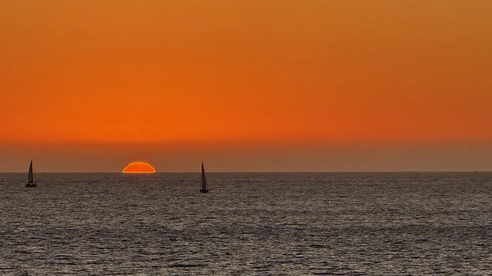 The setting sun glows orange above the ocean horizon as two sailboats cross the rippling water.
