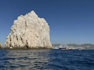 A towering pale rocky cliff rises from choppy blue ocean water beside several boats, with distant mountains along the shore.