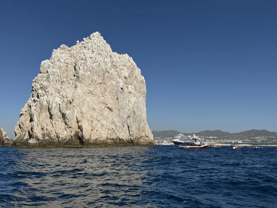 A towering pale rocky cliff rises from choppy blue ocean water beside several boats, with distant mountains along the shore.