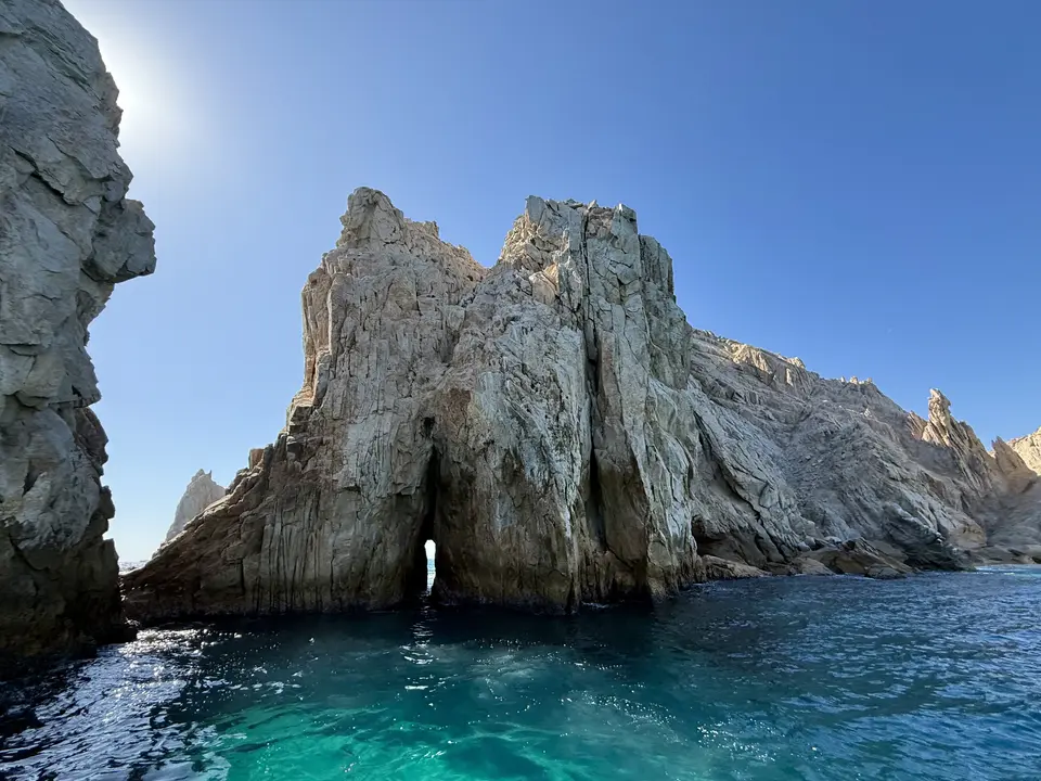 Jagged pale rock cliffs rise from clear turquoise ocean water beneath a cloudless blue sky.