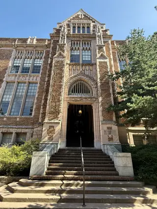 A Gothic-style brick building features tall mullioned windows, carved stone details, and a recessed arched entrance above a broad staircase.