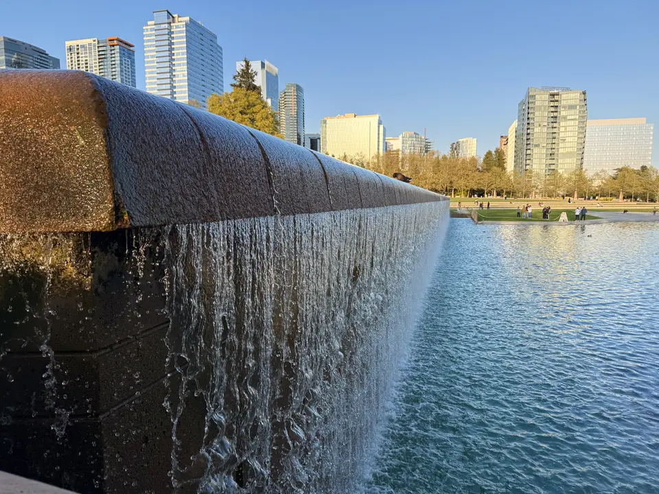 Water spills over the edge of a broad fountain pool with modern high-rise buildings and a grassy park in the background.