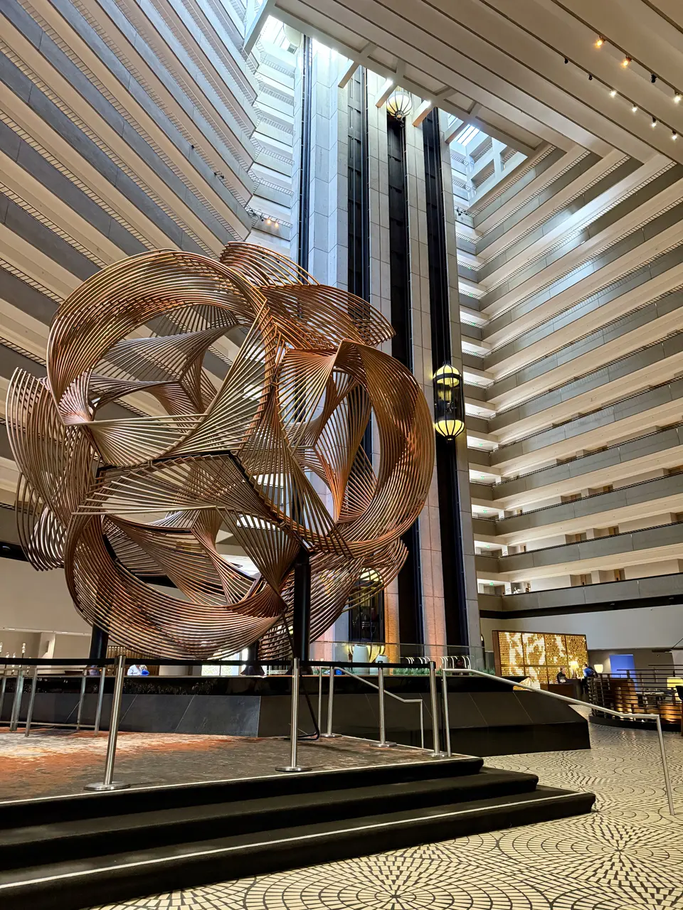 A large abstract metal sculpture stands in the atrium of a multi-story hotel-like interior with balconies and glass elevators.